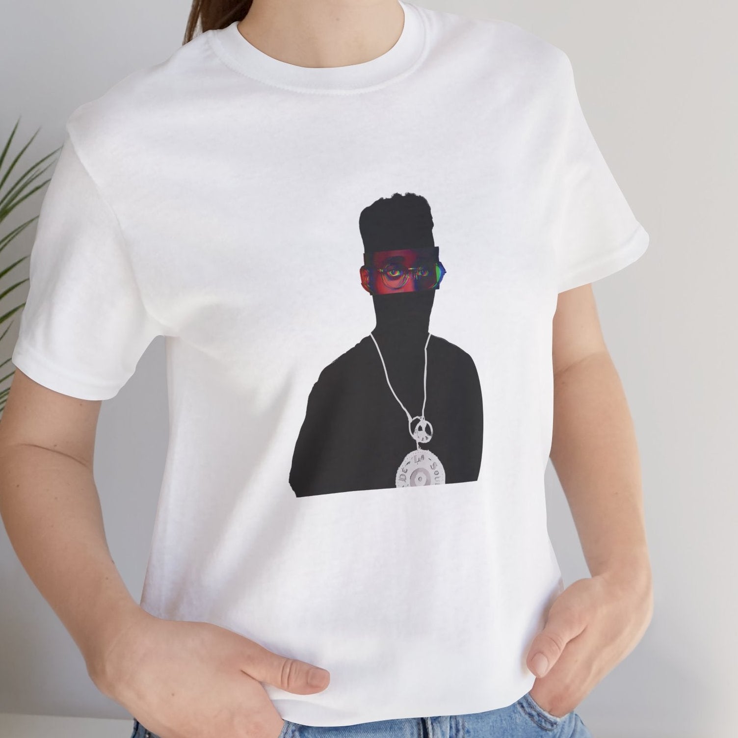 Alternative Hip-Hop music icon graphic t-shirt in white featuring illustrated silhouette portrait design with high-top fade, round glasses, peace medallion and chain - styled with denim jacket in studio setting - IconBandwear music icons collection
